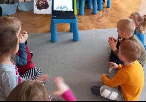 Dzieci siedzą w kółku na dywanie, na wprost laptopa.