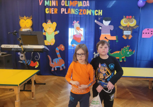 Dwóch chłopców stoi na tle dekoracji V Mini Olimpiady Gier Planszowych.
