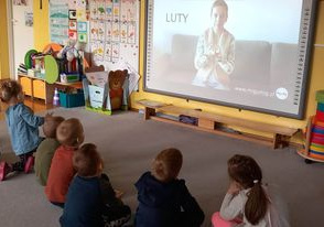 Dzieci siedzą na dywanie przed tablicą multimedialną, uczą się języka migowego.