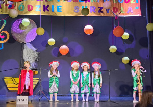 Grupa dzieci stoi na scenie, przed mikrofonami i śpiewa piosenkę o Łodzi.