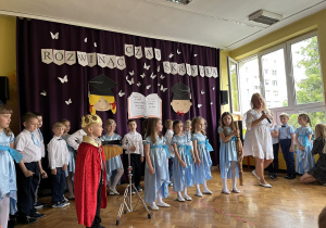 Dzieci stoją w sali, uczestniczą w uroczystości zakończenia roku przedszkolnego. Śpiewają piosenki, tańczą i recytują wiersze.
