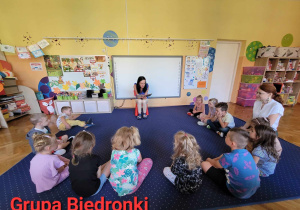 Dzieci z grupy Biedronki siedzą na dywanie w kole, słuchają głośno czytającej bajki zaproszonej Pani.
