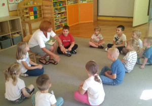 Dzieci z grupy Liski siedzą na dywanie w kole, słuchają głośno czytającej bajki nauczycielki.