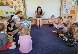 Dzieci z grupy Biedronki siedzą na dywanie w kole, słuchają głośno czytającej bajki zaproszonej Pani.