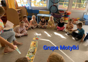 Dzieci z grupy Motylki siedzą na dywanie w kole, słuchają głośno czytającej bajki nauczycielki.