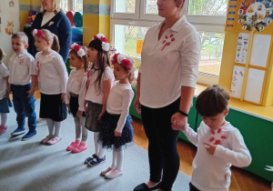 Dzieci stoją w kole, ubrane na galowo, śpiewają hymn Polski