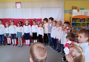 Dzieci stoją w kole, ubrane na galowo, śpiewają hymn Polski