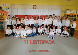 Dzieci stoją i siedzą, ubrane na galowo, na tle flagi Polski i godła Polski.