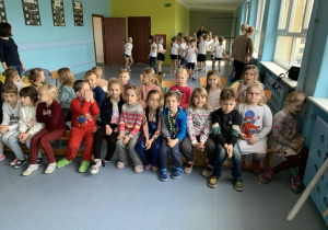 Dzieci siedzą na ławce i krzesełkach w oczekiwaniu na przedstawienie.
