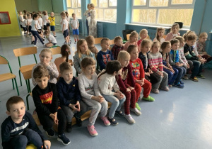 Dzieci siedzą na ławce i krzesełkach w oczekiwaniu na przedstawienie.