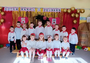 Dzieci stoją i siedzą w grupie na tle dekoracji z okazji Dnia Babci i Dziadka.