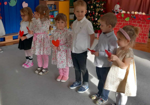 Dzieci stoją w półkolu, trzymając czerwone serca w rękach.