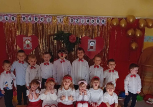 Dzieci stoją i siedzą w grupie na tle dekoracji z okazji Dnia Babci i Dziadka.