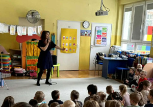 Pani Natalia stoi przed dziećmi siedzącymi przed nią i śpiewa piosenkę.