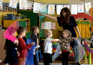 Pięcioro dzieci tańczy z kolorowymi chustami z panią Natalią na środku sali, przed nimi siedzą pozostałe dzieci.