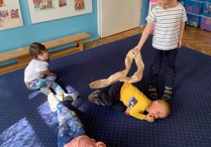 Choreoterapia w grupie I. Dzieci leżą na dywanie. Jedno muska je chustą po plecach.