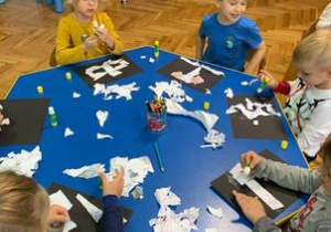 Plastykoterapia w grupie I. Dzieci siedzą przy stoliku, wydzierają z białego kartonu wzory i naklejają na czarny karton.