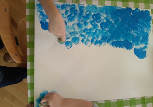 Plastykoterapia w grupie 4. Dwoje dzieci stempluje duży karton gąbką maczaną w niebieskiej farbie.