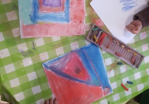 Plastykoterapia w grupie 4. Kilkoro dzieci siedzi przy stoliku i suchymi pastelami rysuje na kwadratowych kartonach.