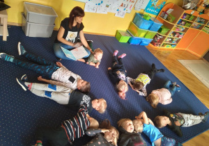 Biblioterapia w grupie 4. Dzieci leżą na plecach w kole z głową skierowaną do środka koła. Słuchają bajki relaksacyjnej czytanej przez nauczyciela.