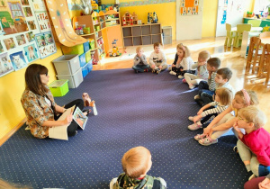 Biblioterapia w grupie 4. Dzieci siedzą w siadzie skrzyżnym przed nauczycielką. Słuchają czytanej przez nią bajki. Podczas spotkania wykorzystywane są ilustracje oraz pacynki: wiewiórka i sowa.