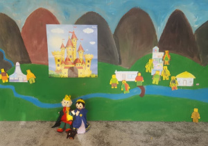 Teatroterapia w grupie 4. Na zdjęciu widać pracę zbiorową, która powstała podczas kilku zajęć. Zdjęcie prezentuje namalowaną farbami scenerię królestwa, pacynki króla i królowej oraz ich psa, a także pacynki wykonane przez dzieci - mieszkańcy królestwa oraz ich domy.