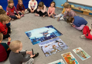 Biblioterapia w grupie 1. Dzieci siedzą w kole na dywanie. Po środku rozłożone są ilustracje przedstawiające zimę i sposoby dokarmiania i dbania o zwierzęta i ptaki podczas zimy.