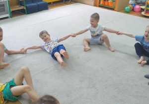 Grupa 5. Grupa dzieci siedzi na dywanie, trzymając się za ręce, przeciągając swoje ciało na prawą i lewą stronę.