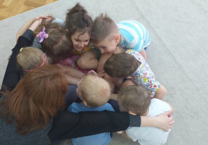 Grupa 5. Grupa dzieci siedzi w kole na dywanie, witając się poprzez przytulenie.
