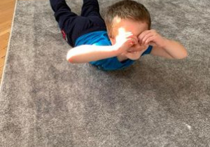 Grupa 1. Chłopiec leży na dywanie, na brzuchu. Unosi nogi do góry, a z rąk przyłożonych do oczu tworzy lornetkę.