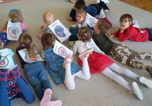 Biblioterapia grupa 5. Dzieci leżą na dywanie z przyklejonymi na plecach obrazkami pokolorowanych muszelek, słuchając "Bajki o Ślimaku i Żółwiu".