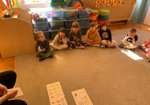 Biblioterapia w grupie 1. Dzieci siedzą na dywanie. Po środku leżą ilustracje do treści opowiadania.