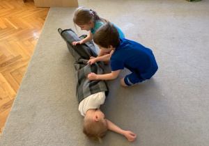 Gr. 1. Dwoje dzieci zawija kolegę w koc turlając go na dywanie.