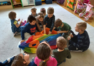 Teatroterapia grupa 5. Dzieci siedzą na dywanie, losując różne zabawki z kolorowego kosza, przykrytego kolorową chustą.