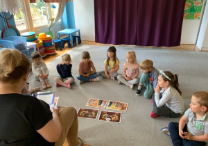 Gr.1. Biblioterapia. Dzieci siedzą na dywanie w kole. Nauczycielka czyta opowiadanie, przed dziećmi rozłożone są ilustracje do opowiadania.