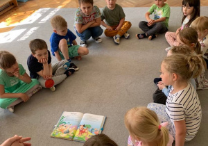 Gr. 1. Biblioterapia. Dzieci siedzą na dywanie wokół ilustracji do opowiadania czytanego przez nauczycielkę.