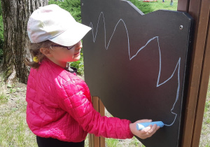 Terapia ręki grupa 2. Na zdjęciu widać dziewczynkę rysującą kredą po tablicy w ogrodzie przedszkolnym.