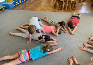 Gr. 1. Dzieci leżą na brzuchu na dywanie tworząc okrąg. Jedno z dzieci przechodzi na czworakach nad leżącymi.