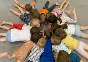 Grupa 1. Dzieci leżą obok siebie na dywanie, na brzuchu. Tworzą koło, nogi rozstawione w kształcie promieni.