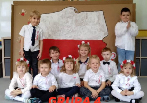 Dzieci z grupy 4 siedzą na tle pracy plastycznej - mapy Polski.