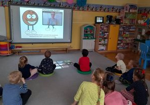 Dzieci siedzą na dywanie przed tablicą multimedialną i uczą się języka migowego.