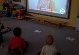 Dzieci siedzą na dywanie przed tablicą multimedialną i uczą się języka migowego.