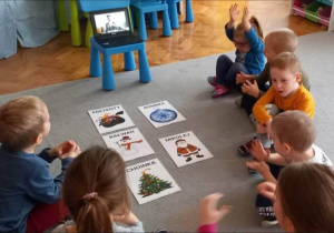 Dzieci siedzą na dywanie, wokół rozłożonych na podłodze obrazków, na wprost laptopa.