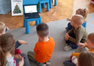 Dzieci siedzą w kółku na dywanie, na wprost laptopa.