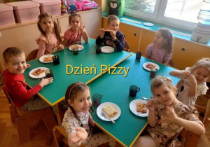 Dzieci siedzą przy stole i jedzą pizzę.