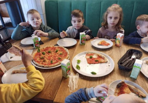 Dzieci siedzą w pizzerii przy stoliku i jedzą pizzę.
