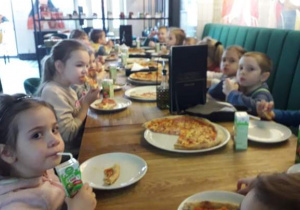 Dzieci siedzą w pizzerii przy stoliku i jedzą pizzę i piją sok.