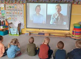 Dzieci siedzą na dywanie przed tablicą multimedialną, uczą się języka migowego.