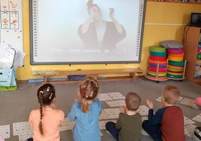 Dzieci siedzą na dywanie przed tablicą multimedialną, uczą się języka migowego.