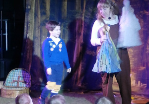 Chłopiec stoi na scenie z aktorką, trzymającą w dłoni kukiełki.